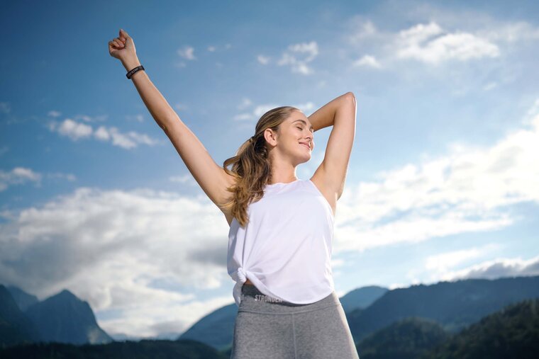 Eine sportlich gekleidete Frau steht unter freiem Himmel und streckt ihre Arme genüsslich von sich weg.